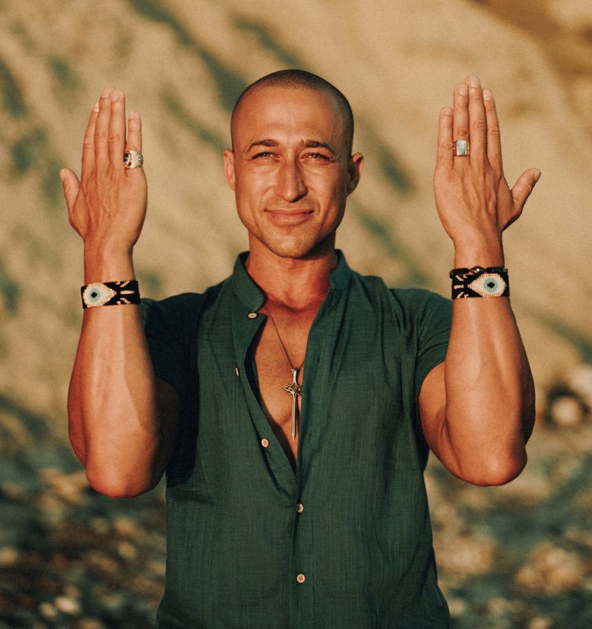 Vuja Deus outdoors in warm light, green shirt, hands raised in prayer, centered and calm.